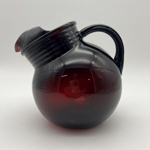 May include: A dark red glass pitcher with a spherical body, a wide spout, and a curved handle. The glass features a ribbed texture around the neck area and a smooth, rounded base. The material is translucent, allowing light to pass through.