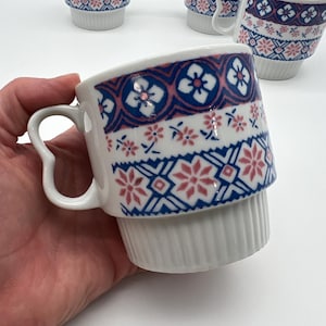 May include: A set of white ceramic mugs featuring a repeating pattern of blue and pink geometric shapes and floral motifs. Each mug has a distinct handle shape and a ribbed base design. The mugs are stackable and feature a smooth, glossy finish.