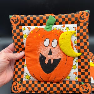May include: A square fabric potholder featuring a central orange pumpkin with a smiling face and a yellow crescent moon. The border is a black and orange checkerboard pattern with pumpkin motifs at the corners, set against a white star-patterned background.