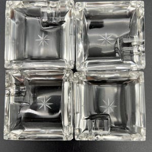 May include: Four clear glass ashtrays. Each square ashtray has a starburst design etched in the center and a notch on one side. The ashtrays are arranged in a square formation.