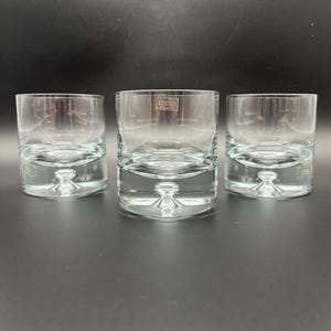 May include: Three clear glass tumblers with thick, heavy bases featuring a single centered air bubble design. Each glass has a straight-sided cylindrical shape and a smooth rim. A small rectangular sticker reading Poland is visible on the center glass.