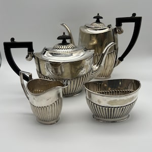 May include: A silver tea set with a teapot, creamer, sugar bowl, and another teapot. The set has black handles and finials. The pieces have a ribbed design and are arranged on a white surface.