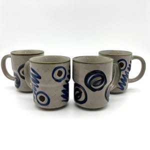 May include: Four ceramic mugs with a speckled, light gray finish. Each mug features a unique design of blue and brown abstract shapes and lines. The rims are trimmed with a green band. The mugs have a handle and a stacked design.