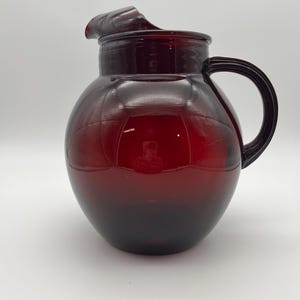 May include: A deep red glass pitcher with a rounded body, a wide circular opening, and a curved handle on the right side. The pitcher features a prominent pouring spout at the top rim and a smooth, translucent surface that reflects light.