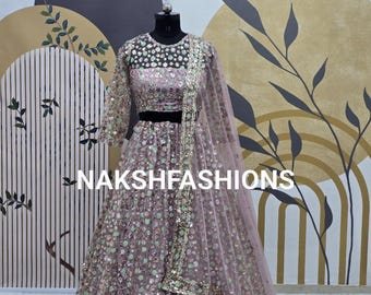 Può includere: Un lehenga rosa chiaro e verde con paillettes, con un top corto e un dupatta abbinati. L'abito è decorato con ricami floreali e paillettes. L'abito è esposto su un manichino su uno sfondo a motivi.