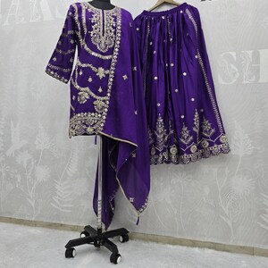 Peut inclure: Un ensemble violet vif comprenant une tunique, une jupe et une &eacute;charpe, tous orn&eacute;s de broderies dor&eacute;es complexes. La tunique a un col rond et des manches trois-quarts. La jupe est longue et fluide. La tenue est pr&eacute;sent&eacute;e sur un mannequin.