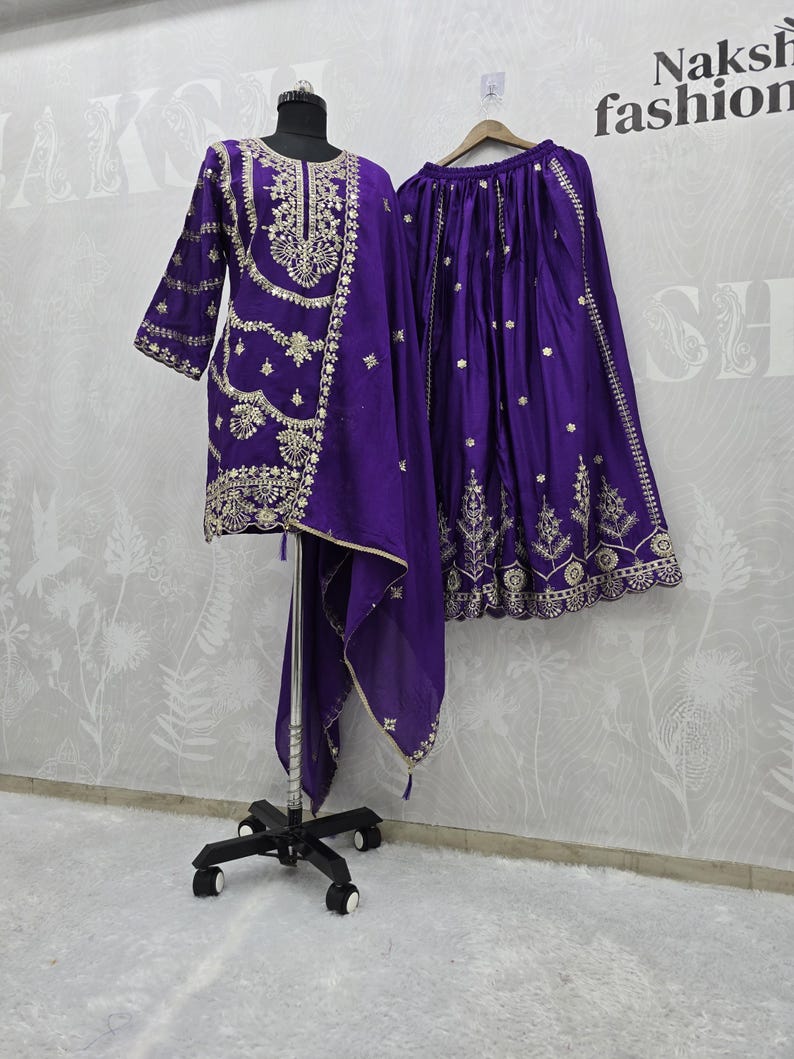 Peut inclure: Un ensemble violet vif comprenant une tunique, une jupe et une &eacute;charpe, tous orn&eacute;s de broderies dor&eacute;es complexes. La tunique a un col rond et des manches trois-quarts. La jupe est longue et fluide. La tenue est pr&eacute;sent&eacute;e sur un mannequin.