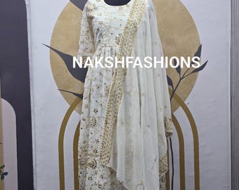 May include: White dress with gold embroidery and a matching sheer dupatta. The dress has a round neckline and long sleeves. The dupatta is draped over the shoulder and has a decorative border.