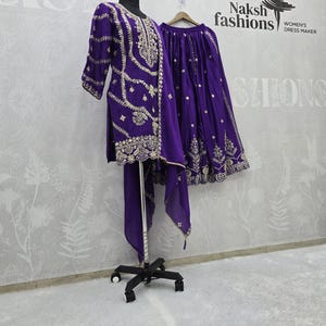 Peut inclure: Une tenue brod&eacute;e violette, comprenant une tunique, une jupe et une &eacute;charpe, pr&eacute;sent&eacute;e sur un mannequin et une cintre. La tenue pr&eacute;sente des broderies dor&eacute;es complexes. Le fond comprend le texte "Naksh fashions WOMEN'S DRESS MAKER".