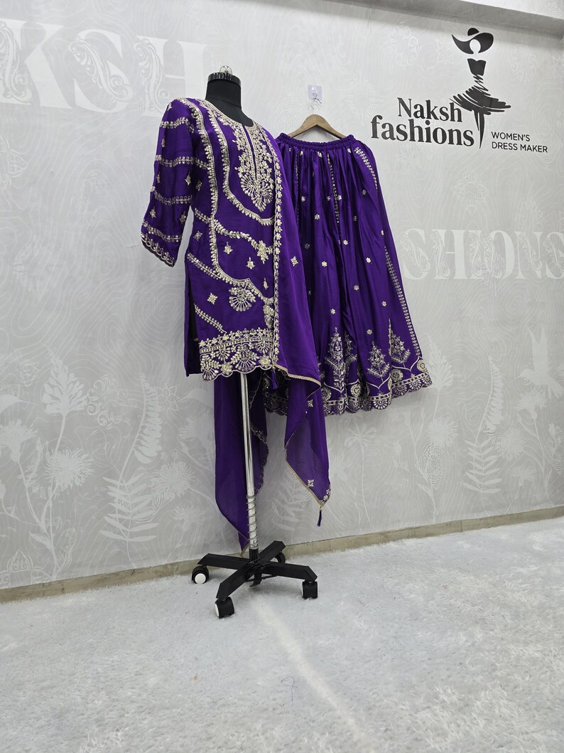 Peut inclure: Une tenue brod&eacute;e violette, comprenant une tunique, une jupe et une &eacute;charpe, pr&eacute;sent&eacute;e sur un mannequin et une cintre. La tenue pr&eacute;sente des broderies dor&eacute;es complexes. Le fond comprend le texte "Naksh fashions WOMEN'S DRESS MAKER".