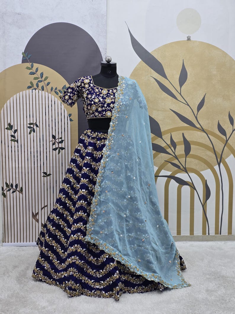 May include: A blue and gold embroidered lehenga with a light blue sheer dupatta. The lehenga has a zig-zag pattern and is decorated with sequins and beads.