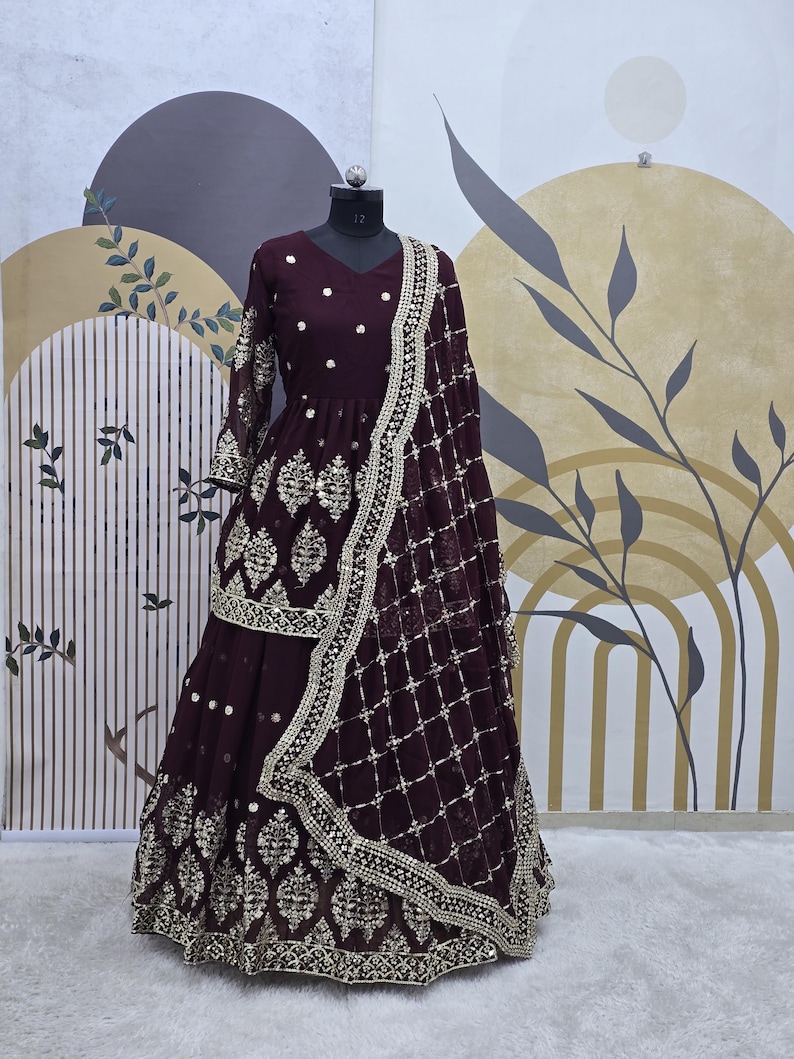 May include: A maroon-colored dress with gold embroidery and sequins. The dress has a long skirt and a fitted bodice. The dress is paired with a matching maroon dupatta with gold embroidery and sequins.