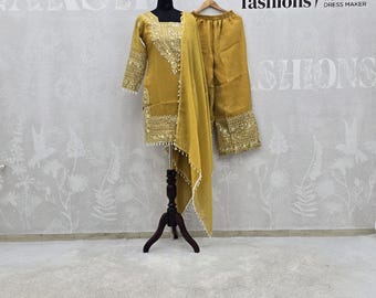 May include: Mustard yellow salwar kameez set with intricate gold embroidery on the sleeves, neckline, and hem. The set includes a matching dupatta and pants. The outfit is displayed on a black dress form. The brand logo "Naksh fashions" is visible.