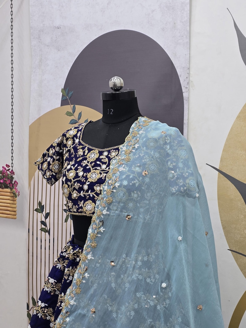 May include: A blue and gold embroidered bridal lehenga with a light blue sheer dupatta. The dupatta has a floral design and is decorated with sequins.