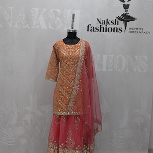 May include: An orange and pink three-piece outfit on a mannequin, featuring a long-sleeved tunic with gold embroidery and mirror work, a matching pink skirt with a wide gold embroidered hem, and a sheer pink dupatta with gold trim and embroidery.