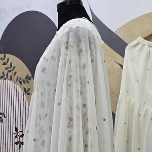 May include: A sheer white fabric with floral embroidery and gold sequins is draped over a mannequin. The fabric has a delicate, airy quality, suggesting it is lightweight. The background features a patterned wall and a glimpse of a matching garment.