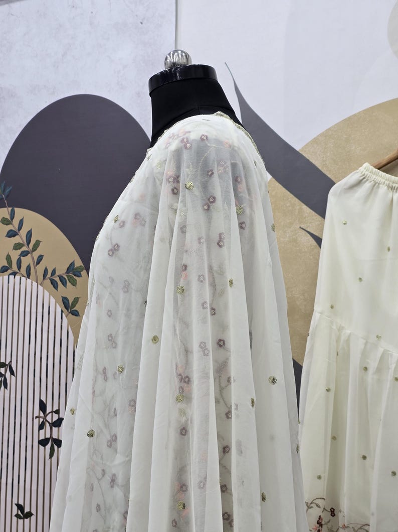 May include: A sheer white fabric with floral embroidery and gold sequins is draped over a mannequin. The fabric has a delicate, airy quality, suggesting it is lightweight. The background features a patterned wall and a glimpse of a matching garment.
