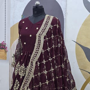May include: A maroon chiffon dress with gold embroidery and sequins. The dress has a V-neckline and a flared skirt. It is paired with a matching maroon net dupatta with gold embroidery and sequins.