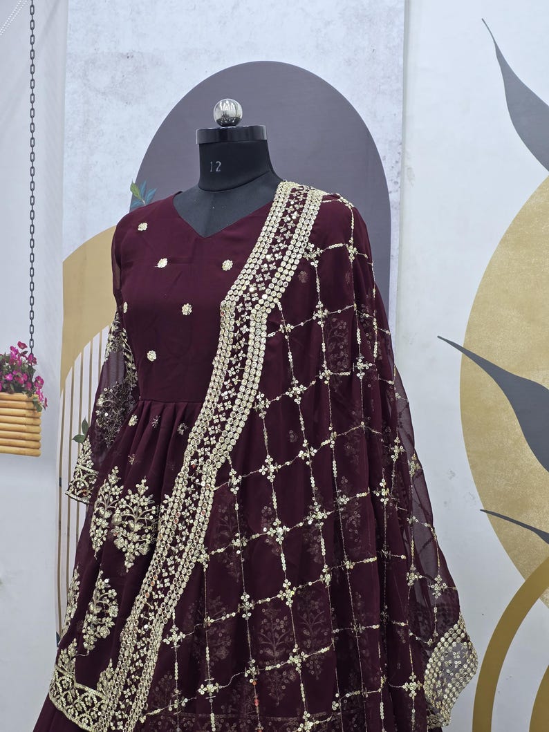 May include: A maroon chiffon dress with gold embroidery and sequins. The dress has a V-neckline and a flared skirt. It is paired with a matching maroon net dupatta with gold embroidery and sequins.