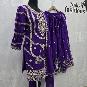 Peut inclure: Une tenue brod&eacute;e violette, comprenant une tunique, un pantalon et une jupe, pr&eacute;sent&eacute;e sur un mannequin. La tenue pr&eacute;sente des broderies dor&eacute;es complexes le long de l'encolure, des manches et de l'ourlet. Le texte "Naksh fashions" est visible en arri&egrave;re-plan.