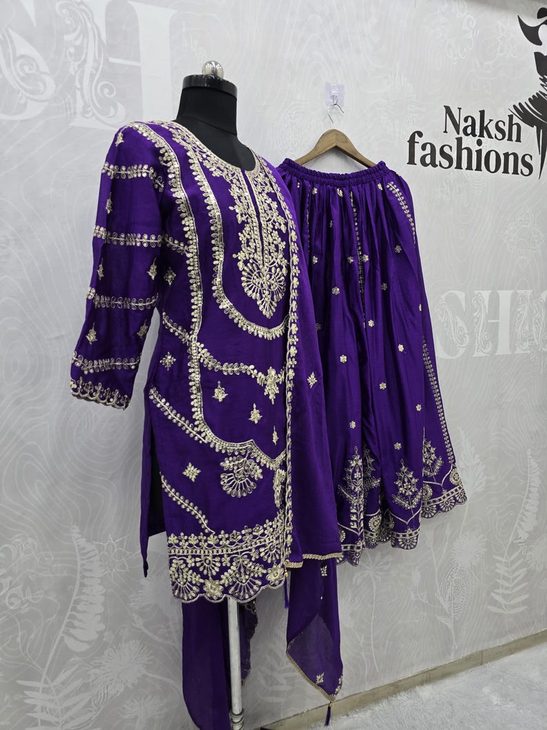Peut inclure: Une tenue brod&eacute;e violette, comprenant une tunique, un pantalon et une jupe, pr&eacute;sent&eacute;e sur un mannequin. La tenue pr&eacute;sente des broderies dor&eacute;es complexes le long de l'encolure, des manches et de l'ourlet. Le texte "Naksh fashions" est visible en arri&egrave;re-plan.