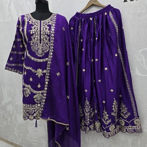 Peut inclure: Ensemble brod&eacute; violet vif, comprenant une tunique &agrave; manches longues, une jupe assortie et une &eacute;charpe transparente. La tunique et la jupe pr&eacute;sentent des broderies dor&eacute;es complexes le long de l'encolure, des manches et de l'ourlet. L'ensemble est pr&eacute;sent&eacute; sur un mannequin et un cintre.