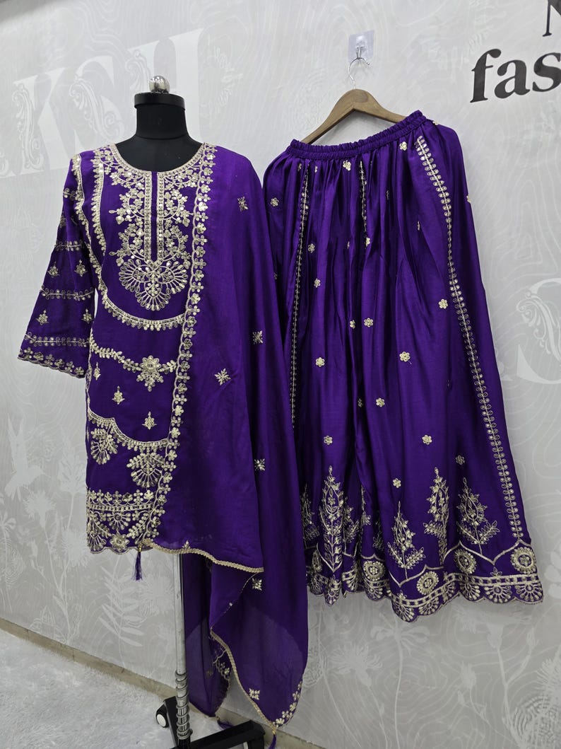 Peut inclure: Ensemble brod&eacute; violet vif, comprenant une tunique &agrave; manches longues, une jupe assortie et une &eacute;charpe transparente. La tunique et la jupe pr&eacute;sentent des broderies dor&eacute;es complexes le long de l'encolure, des manches et de l'ourlet. L'ensemble est pr&eacute;sent&eacute; sur un mannequin et un cintre.