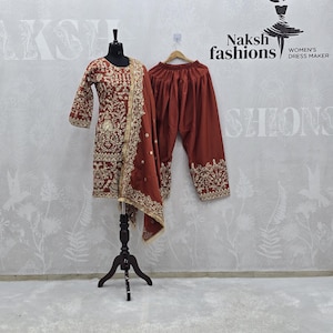 Puede incluir: Un conjunto salwar kameez bordado de color granate con un dupatta a juego. El atuendo se exhibe en un maniquí y una percha. El conjunto presenta intrincados bordados dorados y es de Naksh Fashions.
