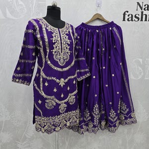 Peut inclure: Ensemble deux pi&egrave;ces violet avec broderies dor&eacute;es. Le haut a des manches longues et un motif d&eacute;taill&eacute; autour de l'encolure et de l'ourlet. La jupe est violette assortie avec une broderie dor&eacute;e similaire. L'ensemble est pr&eacute;sent&eacute; sur un mannequin.