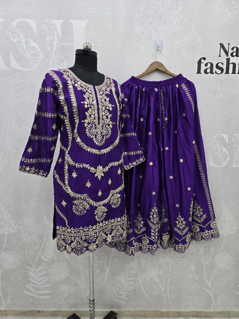 Peut inclure: Ensemble deux pi&egrave;ces violet avec broderies dor&eacute;es. Le haut a des manches longues et un motif d&eacute;taill&eacute; autour de l'encolure et de l'ourlet. La jupe est violette assortie avec une broderie dor&eacute;e similaire. L'ensemble est pr&eacute;sent&eacute; sur un mannequin.