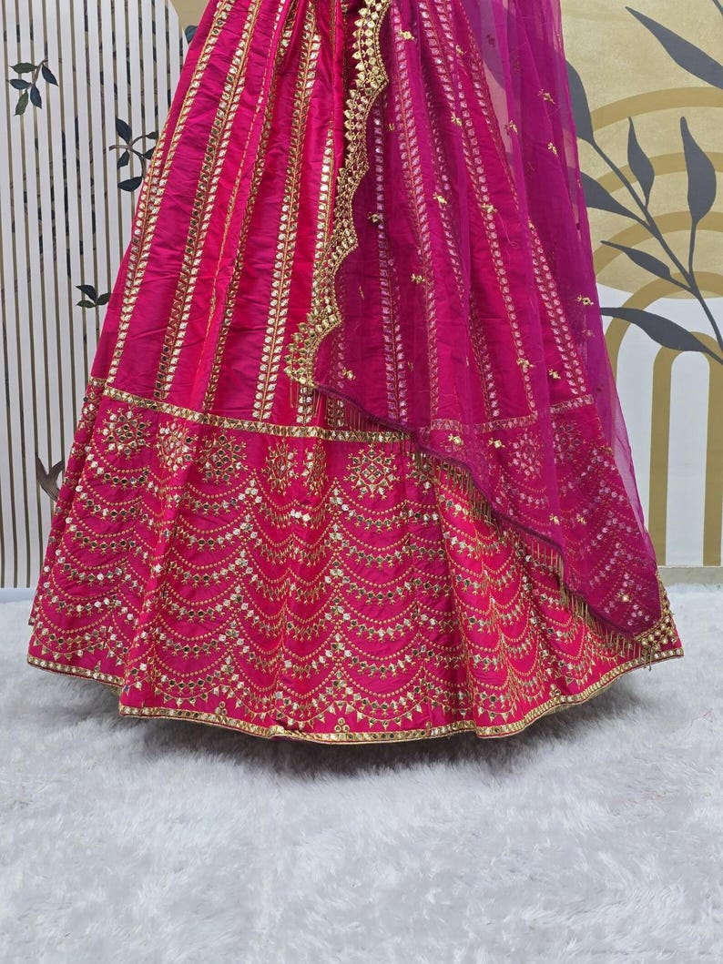 Pode incluir: Uma saia bordada em rosa e dourado com um dupatta transparente a condizer. A saia apresenta trabalhos de espelho intrincados e uma bainha festoneada.