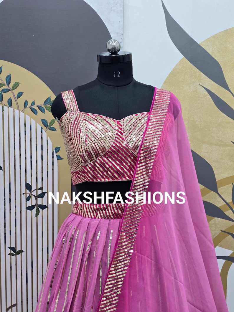 Op de afbeelding: Een roze en gouden sequined lehenga met een bijpassend topje. De outfit wordt getoond op een mannequin tegen een gepatroneerde achtergrond. De tekst "NAKSHFASHIONS" is zichtbaar op de rok.