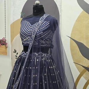 May include: A navy blue lehenga choli set with intricate silver embellishments. The set includes a fitted top with a detailed neckline and a flowing skirt. A sheer, matching dupatta drapes over the shoulders. The outfit is displayed on a mannequin.