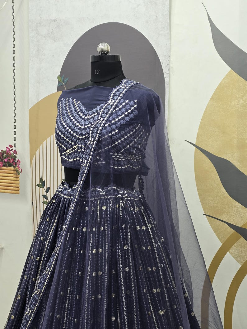 May include: A navy blue lehenga choli set with intricate silver embellishments. The set includes a fitted top with a detailed neckline and a flowing skirt. A sheer, matching dupatta drapes over the shoulders. The outfit is displayed on a mannequin.