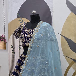 May include: A blue and gold embroidered lehenga with a light blue sheer dupatta. The lehenga has a fitted bodice and a flowing skirt. The dupatta is decorated with sequins and beads.