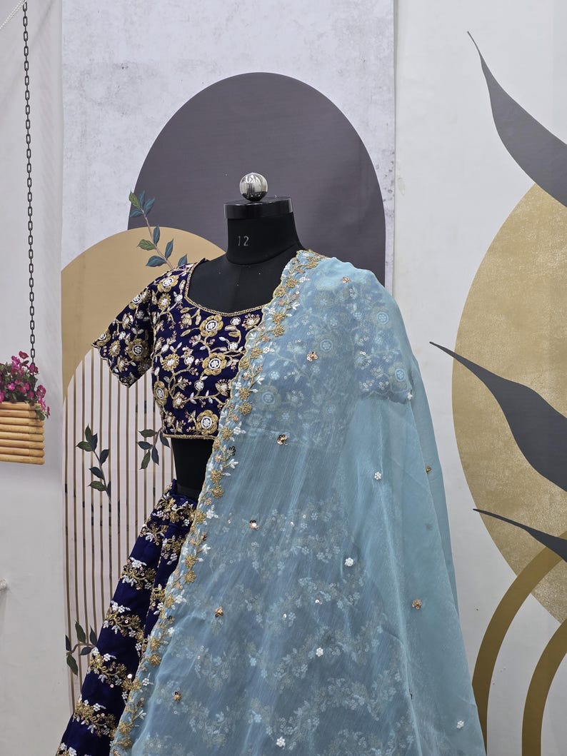 May include: A blue and gold embroidered lehenga with a light blue sheer dupatta. The lehenga has a fitted bodice and a flowing skirt. The dupatta is decorated with sequins and beads.