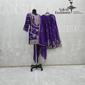Peut inclure: Ensemble violet brod&eacute;, comprenant une tunique, un pantalon et une &eacute;charpe assortie, pr&eacute;sent&eacute; sur un mannequin. Une jupe est suspendue. L'ensemble pr&eacute;sente des broderies dor&eacute;es. La marque "Naksh fashions" est visible.