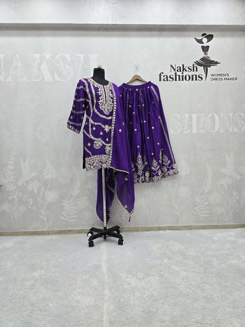 Peut inclure: Ensemble violet brod&eacute;, comprenant une tunique, un pantalon et une &eacute;charpe assortie, pr&eacute;sent&eacute; sur un mannequin. Une jupe est suspendue. L'ensemble pr&eacute;sente des broderies dor&eacute;es. La marque "Naksh fashions" est visible.