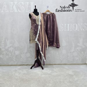 Peut inclure: Ensemble salwar kameez mauve sans manches avec broderies dorées et dupatta assorti avec bordure en dentelle blanche. L'ensemble comprend un pantalon large. L'ensemble est présenté sur un mannequin et un cintre. La marque est Naksh Fashions.