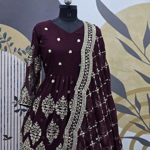 May include: A maroon-colored dress with silver embroidery and embellishments. The dress has a long sleeve and a flared skirt. It is paired with a matching maroon dupatta with silver embroidery.
