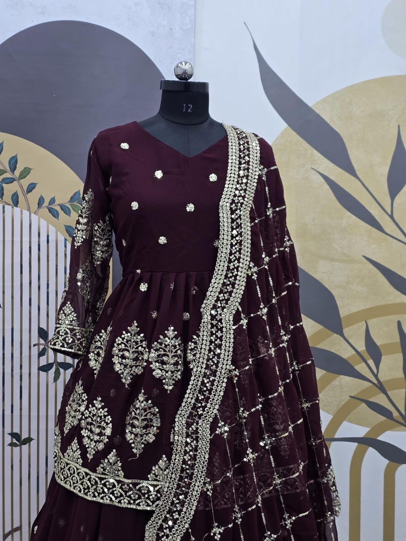 May include: A maroon-colored dress with silver embroidery and embellishments. The dress has a long sleeve and a flared skirt. It is paired with a matching maroon dupatta with silver embroidery.
