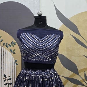 May include: A navy blue two-piece outfit displayed on a mannequin. The top features a geometric pattern of silver and white embellishments. The skirt is pleated and adorned with similar silver accents. The outfit is designed for formal occasions.