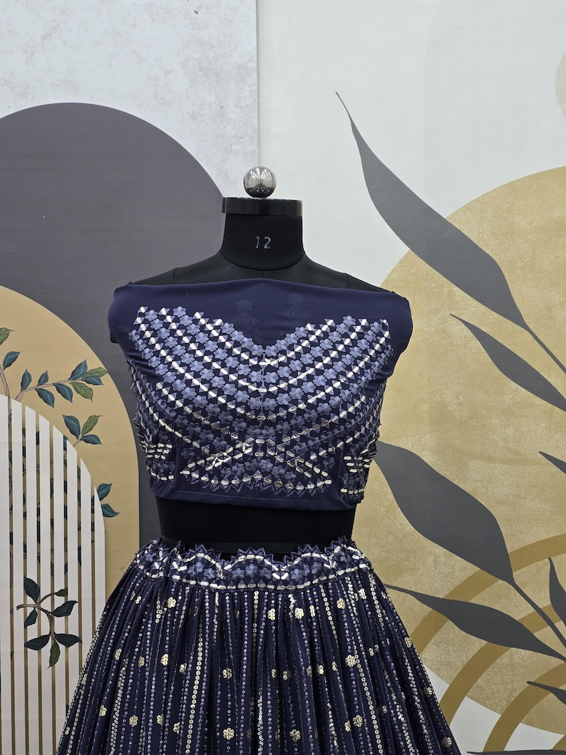 May include: A navy blue two-piece outfit displayed on a mannequin. The top features a geometric pattern of silver and white embellishments. The skirt is pleated and adorned with similar silver accents. The outfit is designed for formal occasions.