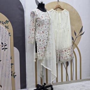 May include: A white embroidered tunic and matching palazzo pants are displayed on a mannequin and hanger. The tunic features floral embroidery with pink and gold accents. The palazzo pants have a similar embroidered hem. The outfit is set against a neutral background.