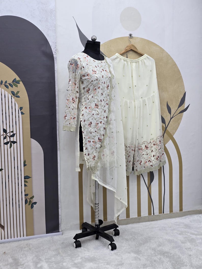 May include: A white embroidered tunic and matching palazzo pants are displayed on a mannequin and hanger. The tunic features floral embroidery with pink and gold accents. The palazzo pants have a similar embroidered hem. The outfit is set against a neutral background.
