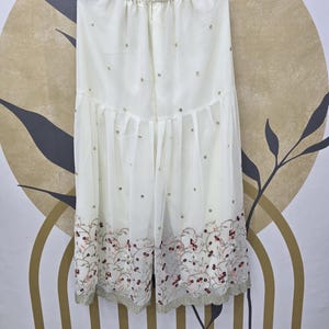 May include: Off-white palazzo pants with an elastic waistband, adorned with scattered sequins and a floral embroidered hemline. The pants are made of a sheer fabric and hang from a wooden hanger.