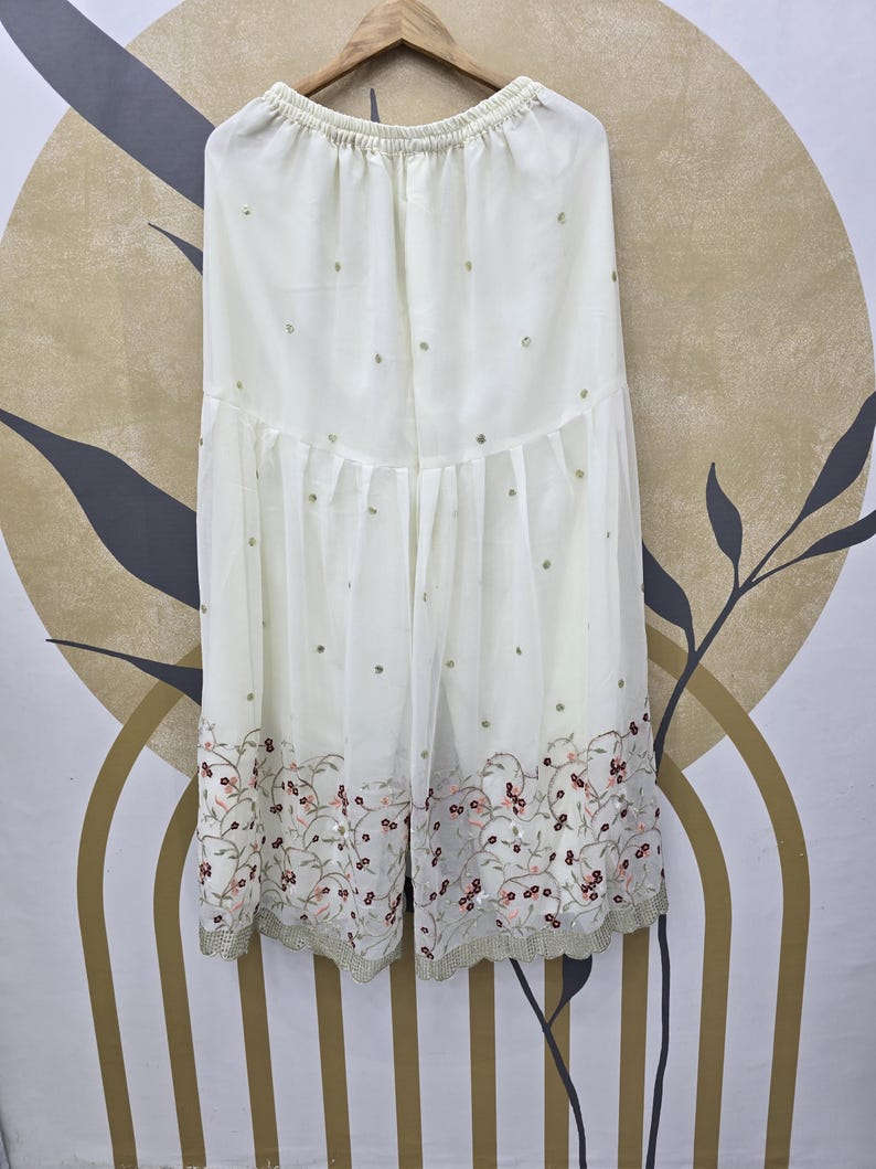 May include: Off-white palazzo pants with an elastic waistband, adorned with scattered sequins and a floral embroidered hemline. The pants are made of a sheer fabric and hang from a wooden hanger.