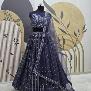 May include: A navy blue lehenga choli set displayed on a mannequin. The set includes a fitted, embellished top, a flowing skirt with vertical stripes and silver dot accents, and a sheer dupatta. The outfit is designed for formal occasions.