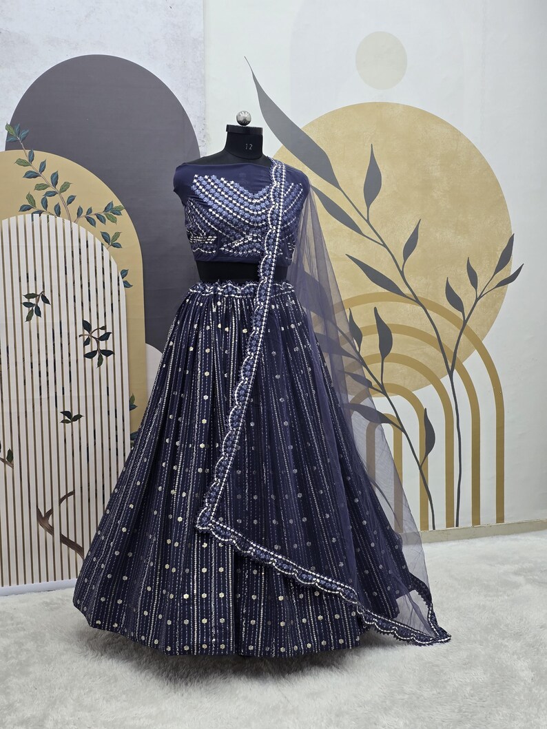 May include: A navy blue lehenga choli set displayed on a mannequin. The set includes a fitted, embellished top, a flowing skirt with vertical stripes and silver dot accents, and a sheer dupatta. The outfit is designed for formal occasions.