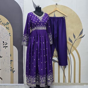Puede incluir: Conjunto morado de vestido de manga larga y pantalón a juego. El vestido presenta intrincados bordados plateados alrededor del cuello, las mangas y el dobladillo. El conjunto se exhibe en un maniquí negro sobre un fondo neutro.